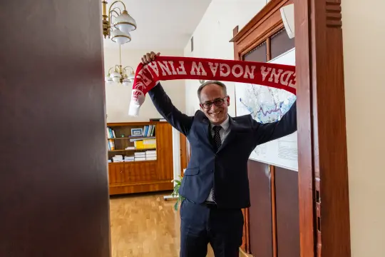 Mężczyzna w garniturze trzyma szalik z napisem w rękach, uśmiechając się.
