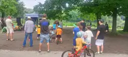 Grupa dzieci i dorosłych na wydarzeniu plenerowym nad jeziorem, z rowerami i sprzętem sportowym.
