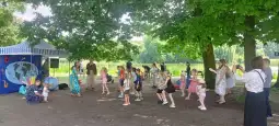 Dzieci tańczą w parku pod drzewami, w tle scena z dekoracjami.