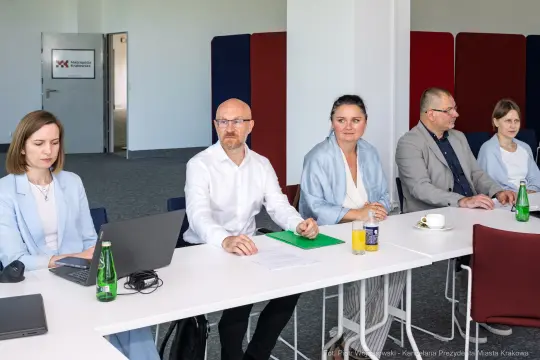 Grupa osób siedzących przy stole w sali konferencyjnej, z laptopami i napojami.