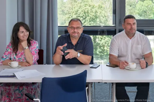 Trzy osoby siedzą przy stole, każda z filiżanką, w tle okna z widokiem na zieleń.