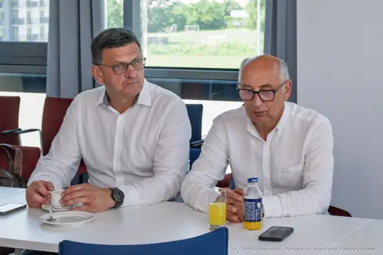 Dwóch mężczyzn w białych koszulach siedzi przy stole, jeden pije sok, drugi kawę.
