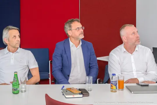 Trzech mężczyzn siedzących przy stole, skupionych na prezentacji. Na stole napoje i notatki.