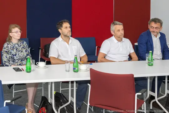 Cztery osoby siedzące przy stole, w tle butelki wody i filiżanka.