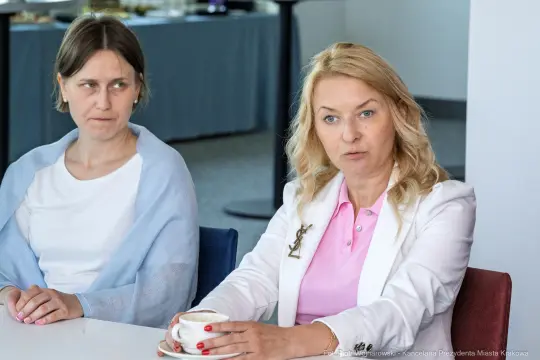 Kobieta w białej marynarce rozmawia przy stole, obok siedzi inna kobieta.