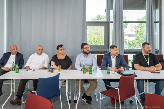 Grupa osób siedzących przy stole, w tle okna i zielone otoczenie.
