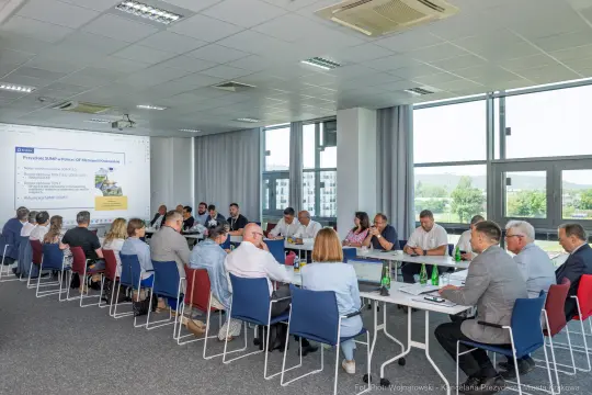 Spotkanie w sali konferencyjnej z uczestnikami przy stole i prezentacją na ekranie.
