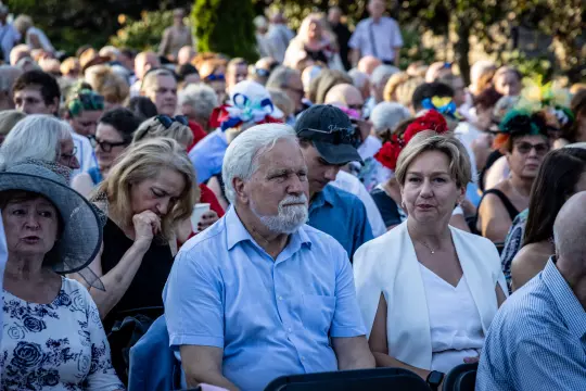 Tłum ludzi siedzących na wydarzeniu, wśród nich starszy mężczyzna i kobieta w białej bluzce.