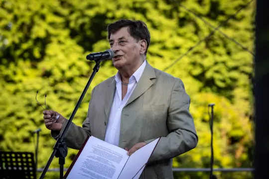 Mężczyzna w jasnym garniturze przemawia do mikrofonu, trzymając kartki z tekstem.