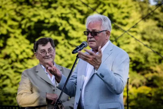 Dwaj mężczyźni przemawiają na scenie, jeden z mikrofonem, w tle zieleń drzew.