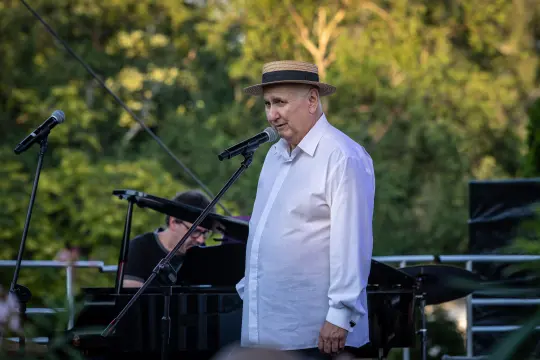 Mężczyzna w kapeluszu śpiewa do mikrofonu, w tle pianista i zieleń.