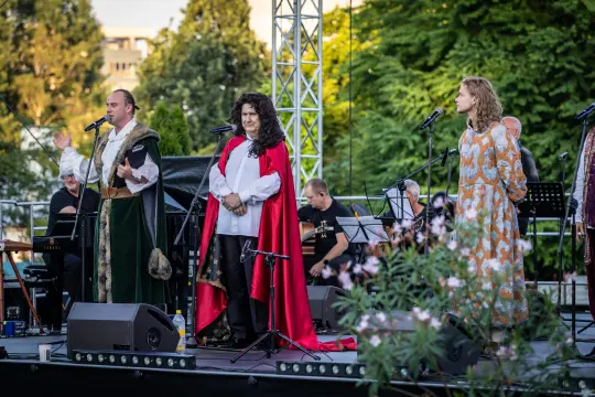 Występ artystów na scenie, w strojach historycznych, z mikrofonami i instrumentami w tle.