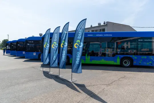 Stojące flagi i autobusy w kolorze niebieskim na tle budynku.