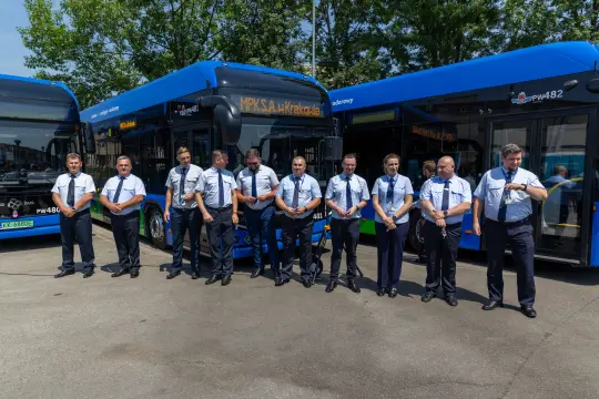 Grupa kierowców autobusów w uniformach, stojących obok pojazdów komunikacji miejskiej.