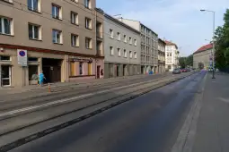Ulica z torami tramwajowymi, budynki mieszkalne i kilka osób na chodniku.