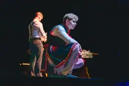 Mężczyzna w kolorowym stroju teatralnym, skupiony na scenie, obok drugi mężczyzna w eleganckim ubraniu.