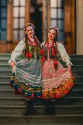 Dwie kobiety w tradycyjnych polskich strojach ludowych z kwiatowymi wiankami na głowach.