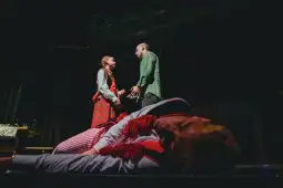 Scena teatralna z dwiema postaciami w dialogu, jedna leży na podłodze.