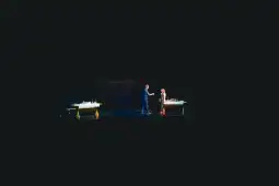 Dwie postacie na scenie, przy stołach z przedmiotami, w interakcji ze sobą.