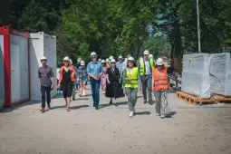 Grupa osób w kaskach i kamizelkach odblaskowych spaceruje po terenie budowy.