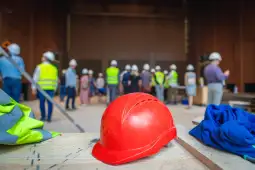 Grupa pracowników budowlanych w kaskach na tle hali. Na pierwszym planie czerwony kask.