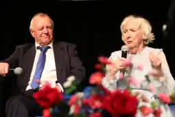 Dwoje starszych ludzi rozmawia na scenie, trzymając mikrofony, w tle kwiaty.