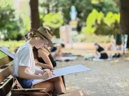Młoda artystka rysująca na ławce w parku, w otoczeniu innych osób.