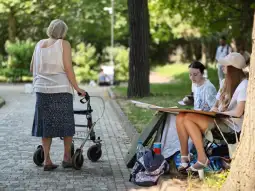 Starsza kobieta z chodzikiem przechodzi obok grupy młodych ludzi siedzących przy stole.
