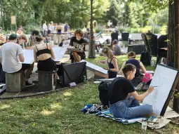 Grupa osób rysujących w parku, siedzących na trawie i kamieniach.