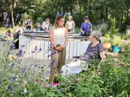 Kobieta rozmawia z seniorką w ogrodzie, w tle inne osoby zajmujące się pracami ogrodowymi.