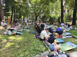 Grupa osób malujących obrazy w parku, otoczona drzewami i zielenią.