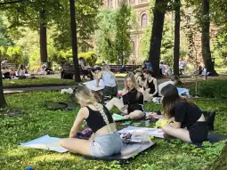 Grupa osób rysujących na trawie w parku, otoczona zielenią i drzewami.