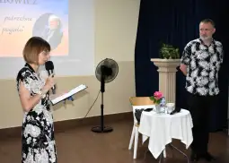 Kobieta przemawia do mężczyzny, obok stół z kwiatami i napojami, w tle prezentacja.
