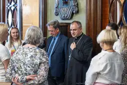 Grupa osób rozmawia w eleganckim wnętrzu, wśród nich duchowny i mężczyzna w garniturze.