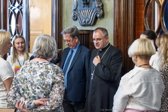 Grupa osób rozmawia w eleganckim wnętrzu, wśród nich duchowny i mężczyzna w garniturze.