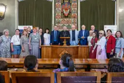 Grupa ludzi w sali, w tle herb miasta, podczas uroczystości lub spotkania.