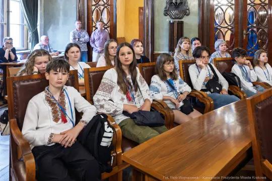 Grupa dzieci w tradycyjnych strojach, siedzących w sali obrad.