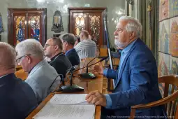 Mężczyzna przemawia w sali obrad, w tle siedzą inni uczestnicy spotkania.