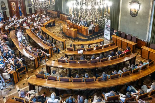 Sala obrad z uczestnikami, stołem prezydialnym i dużym żyrandolem.