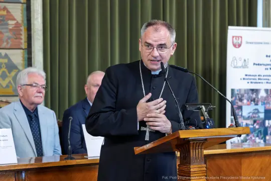 Ksiądz biskup przemawia podczas konferencji, w tle siedzą inni uczestnicy.