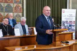 Mężczyzna w garniturze przemawia podczas uroczystości w sali obrad. W tle siedzą inni uczestnicy.