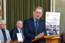 Mężczyzna przemawia podczas wydarzenia w sali, w tle siedzą dwaj inni mężczyźni.