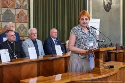 Kobieta przemawia przy mównicy, w tle siedzą mężczyźni w garniturach i sutannach.
