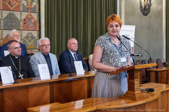Kobieta przemawia przy mównicy, w tle siedzą mężczyźni w garniturach i sutannach.