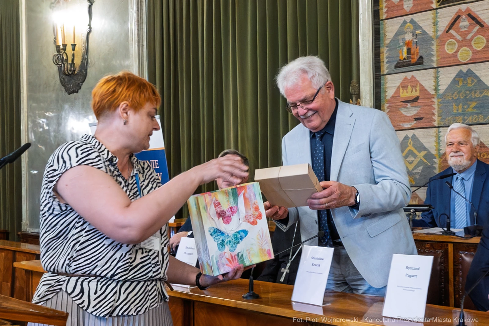 Kobieta wręcza mężczyźnie prezent w sali obrad, w tle widoczni inni uczestnicy spotkania.  Autor: Piotr Wojnarowski