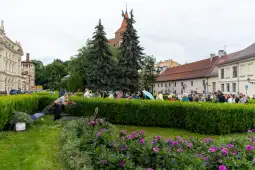 Grupa ludzi w parku, przycięte żywopłoty i kwiaty w tle.