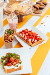 Wafle z bitą śmietaną, truskawkami i sosem, obok napój owocowy i inne słodkości.