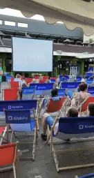 Ludzie siedzą na leżakach, oglądają film na dużym ekranie w plenerze.