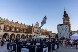 Artystka na szczudłach w białej sukni w centrum Krakowa, tłum ludzi i budynki w tle.