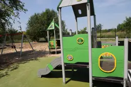 Plac zabaw z zielonymi elementami, zjeżdżalnią i huśtawkami w tle.